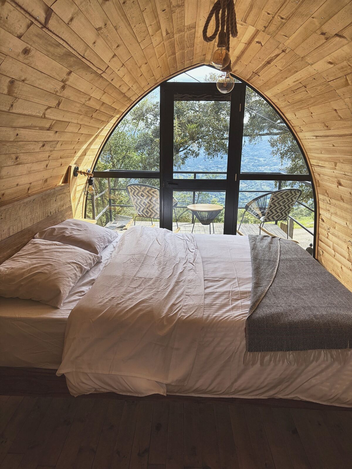 Cama con ropa blanca y manta gris frente al ventanal en arco de la cabaña Alma, con la terraza y el bosque al fondo
